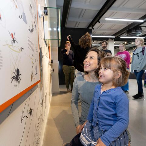 Mutter hockt mit Kind auf dem Schoß an einer Ausstellungstafel im Natur-Erlebniszentrum und sieht sich diese an