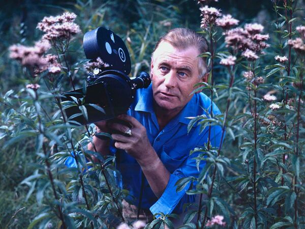 Heinz Sielmann steht in einer Blumenwiese mit einer Kamera in der Hand.