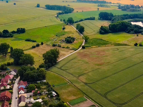 Aufsicht auf Häser und Straßen einer kleinen Ortschaft. Großteil des Bildes ist von landwirtschaftlichen Feldern ausgefüllt. Ein mit Bäumen und Büschen gesäumter See ist am rechten Bildrand zu sehen.gesäumter