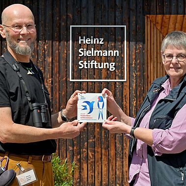 NABU Plakette für Gut Herbigshagen Zwei Personen stehen mit einer Auszeichnung für ein schwalbenfreundliches Haus in den Händen vor einer Holzwand